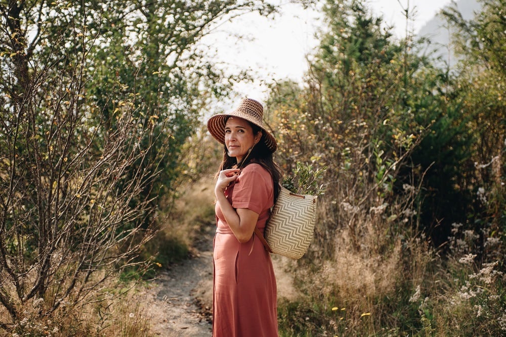 Forager Leigh Joseph wearing a woven hat and backpack as she stands in front of wild plants