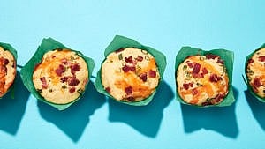 Three cheddar and bacon cornmeal muffins in turquoise wrappers, on blue background.