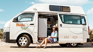 Woman sitting out the side of large white van with blue sky and dirt ground.