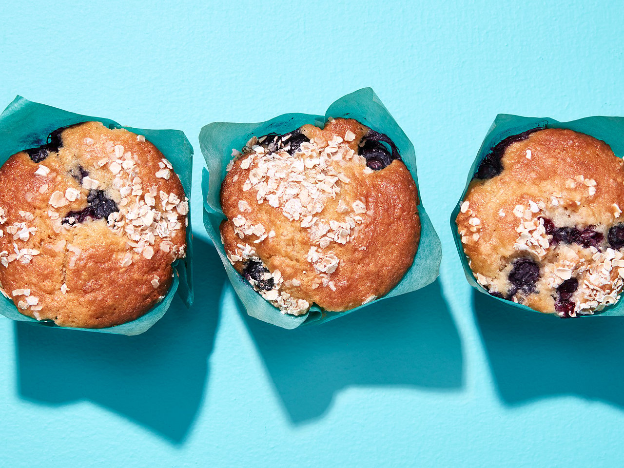 Three blueberry muffins with blue wrapper, on blue background.