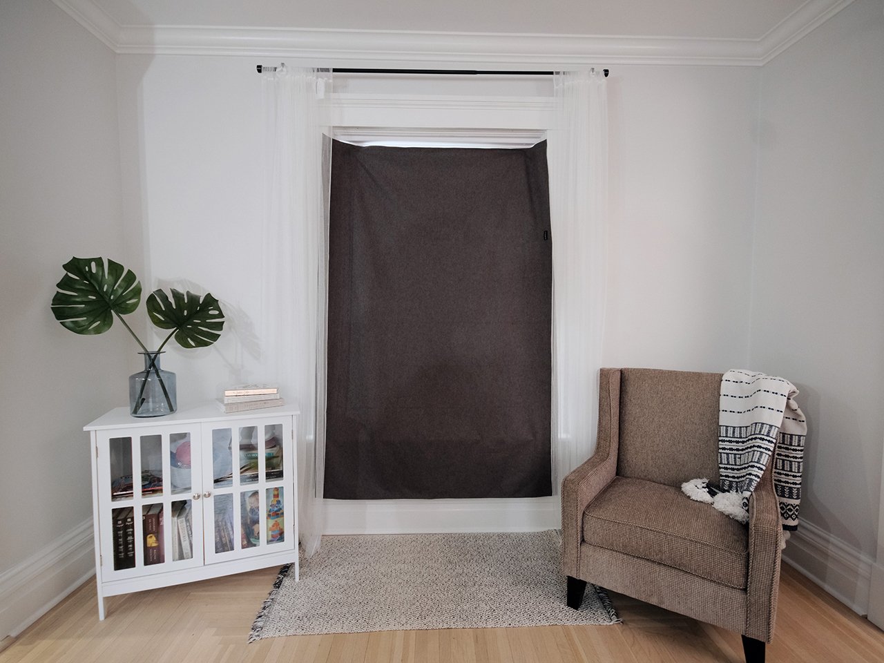 Blackout curtain in front of windor in white room with a rown chair and small white bookshelf.