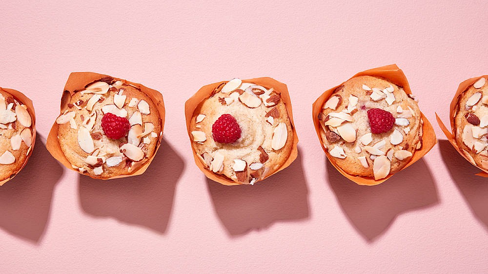 Three gluten-free raspberry and almond muffins in an orange wrapper on pink background.