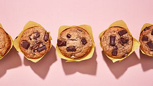 Three vegan banana chocolate chunk muffins in yellow wrapper on a pink background
