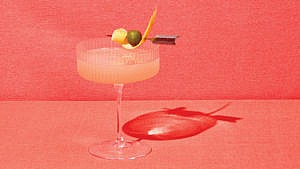 A peach-coloured cocktail with an orange peel and olive on a skewer.