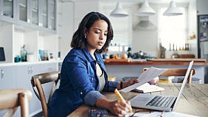 Woman sits at a kitchen dining table in front of a laptop with papers and a pencil in hand.