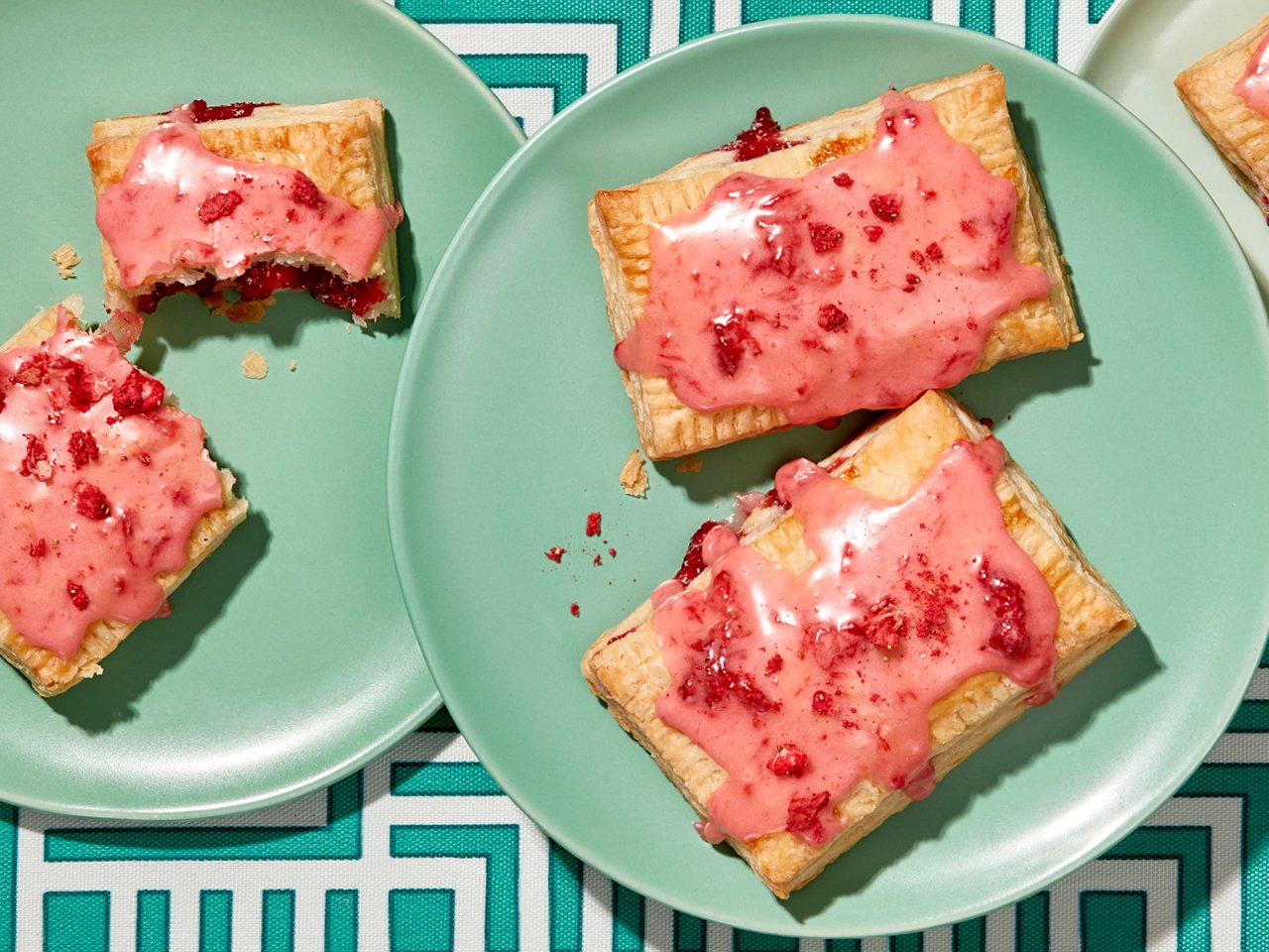 Two pink glazed strawberry handpies on two turquoise plates on a trellis-patterned tablecloth
