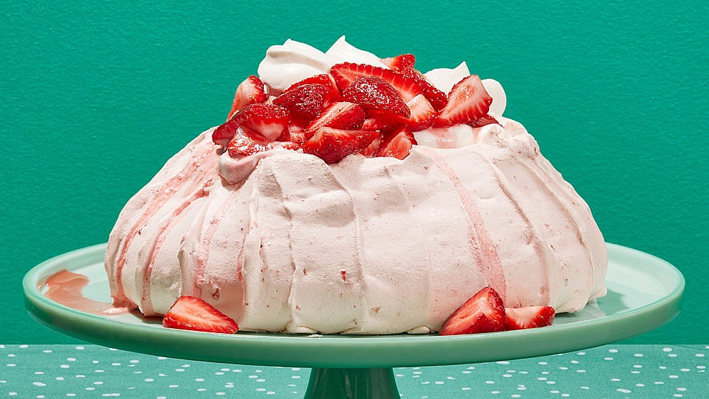 A pink strawberry pavlova topped with fresh strawberries and whipped cream on a turquoise cake stand.