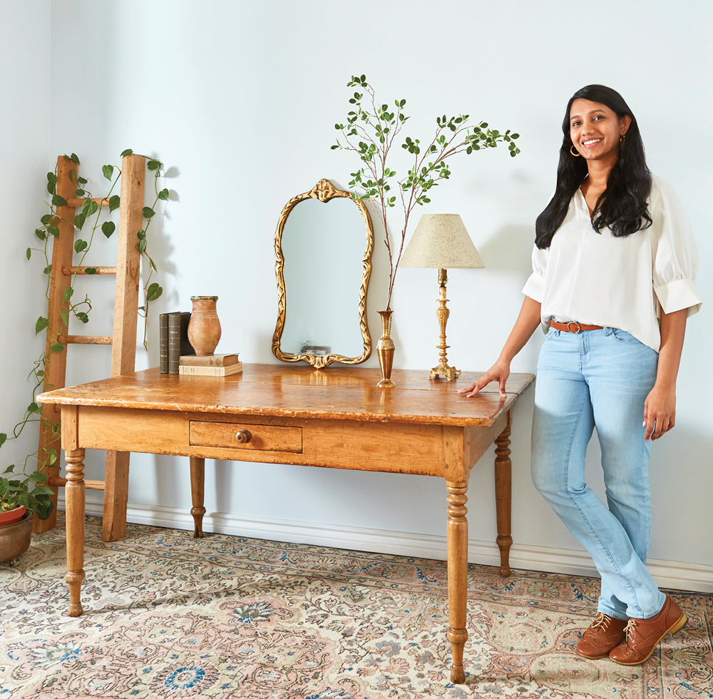 A woman in blue jeans and a white top stands with her hand resting on a wooden table with various lamps , plants and books on it.