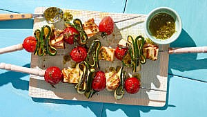 cherry tomato, zucchini and halloumi skewers with a bowl of pesto drizzle on a white gridded rectangular plate on a blue table