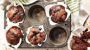 A six-slot muffin tin with four chocolate-zucchini muffins in wrappers in the tin, and one to the side, and two empty slots, on a green and white striped linen tablecloth