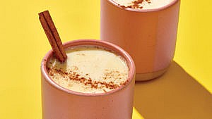 Two cream-coloured drinks in brown mugs against a yellow background with sprinkling of cinnamon on top and a cinnamon stick sticking out of the drink halfway