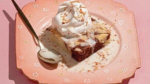 marble cake with whipped cream on top and liquid around the base of the cake, placed in a pink square plate with a metal spoon on the side of the plate.