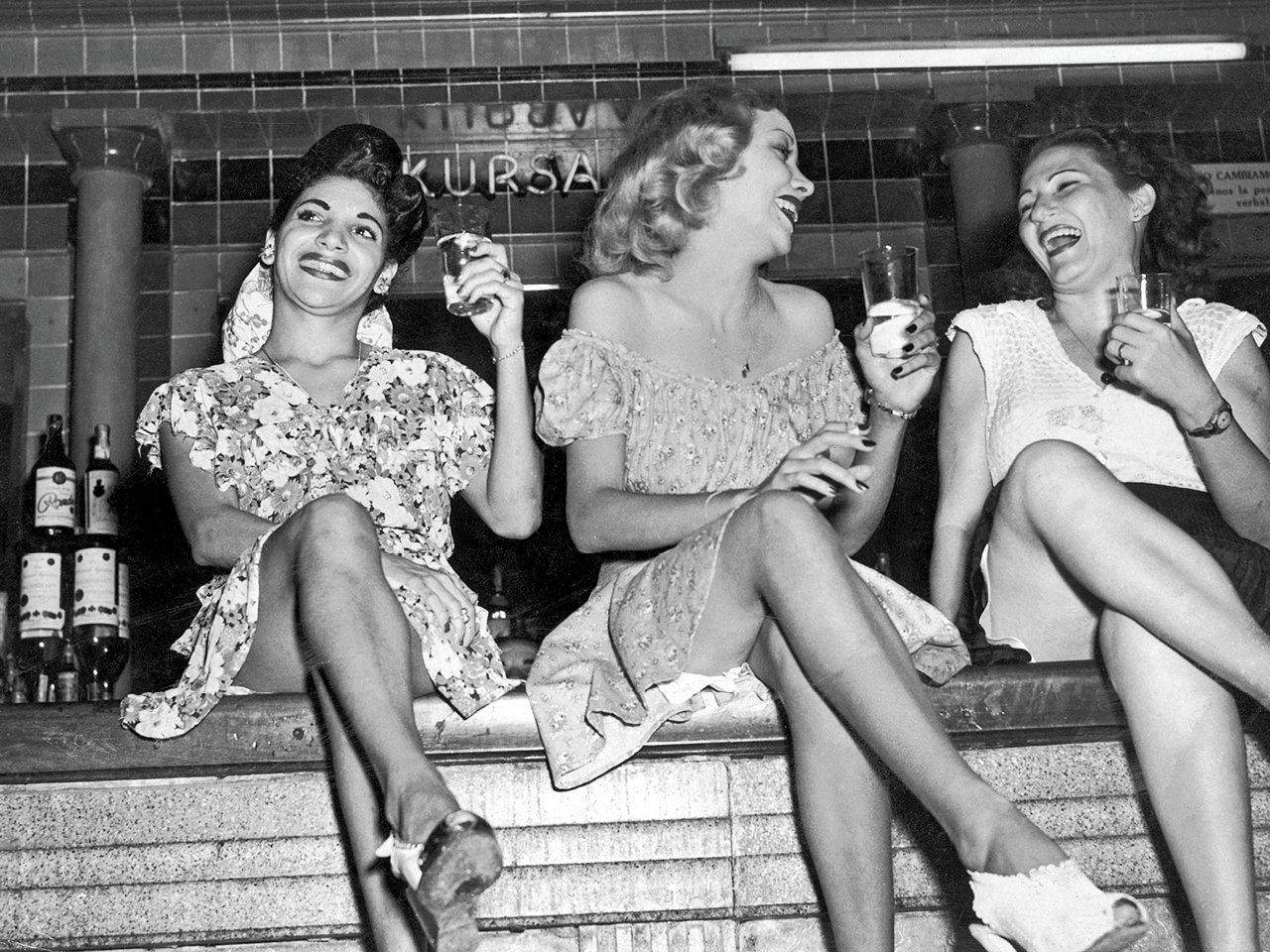 A black and white photo of three young women in dresses, sitting on a bar, holding cocktails and having a great time