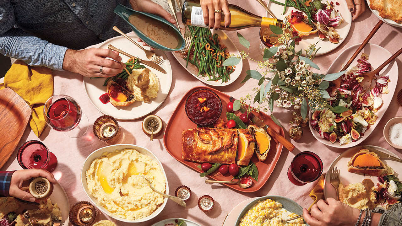 An array of Thanksgiving dishes on a pink table with hands reaching into the frame to eat, pour wine