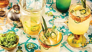 Four spicy pineapple margaritas in different glasses, three rimmed with salt, one with japan peppers garnish, beside a bottle of ginger beer and tequila and alongside a plate of pineapples