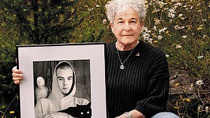 a woman with a turtleneck sweater sits in the woods with a dog at her feet and a black and white photo of her son