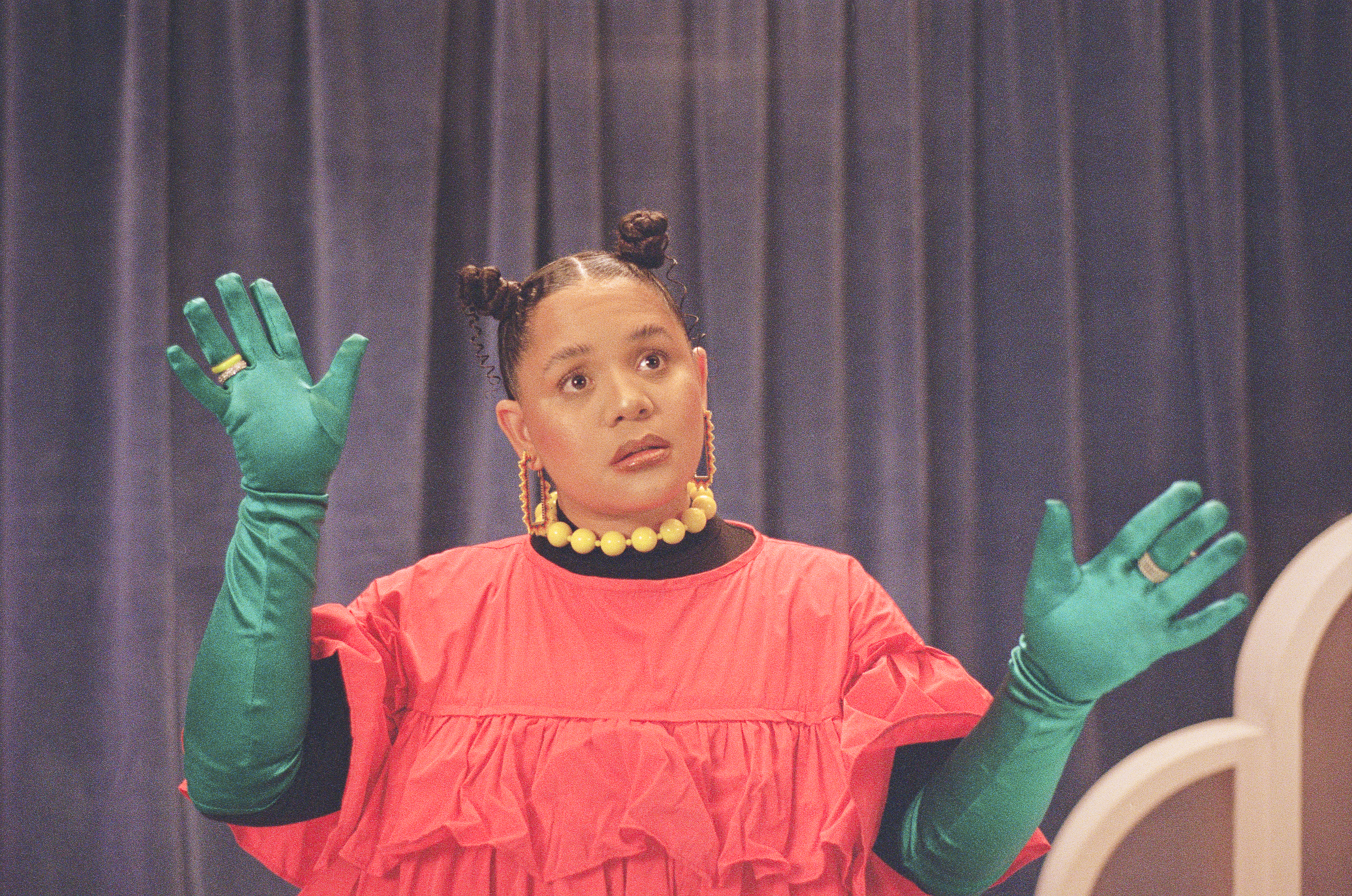 A woman acting on a colourful set stage, wearing a red dress, a bright yellow beaded necklace, blue gloves, with her hair tied into two tight bun knots.