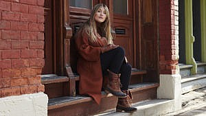 Woman with long blonde hair sitting outside on steps wearing a brown coat, black pants and brown winter boots.