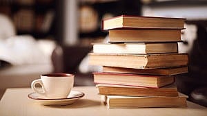 A stack of books on a wood table next to a cup in a saucer