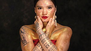 Arsaniq Deer poses with her traditional tattoos on her face and arms, covered in gold glitter and looking ahead at the camera.