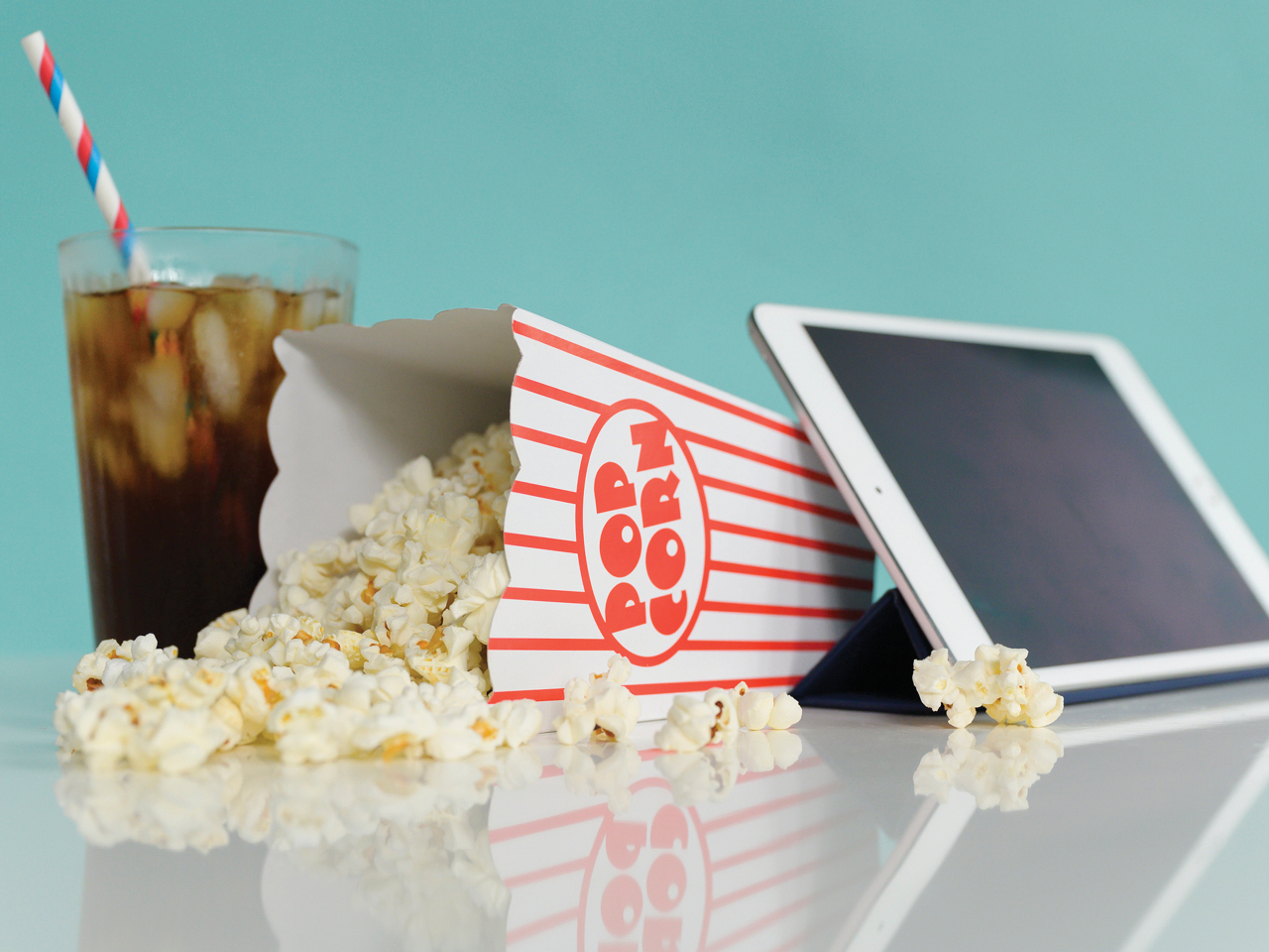 A glass of soda, a tipped over carton of popcorn, and an Ipad are displayed next to each other on a table