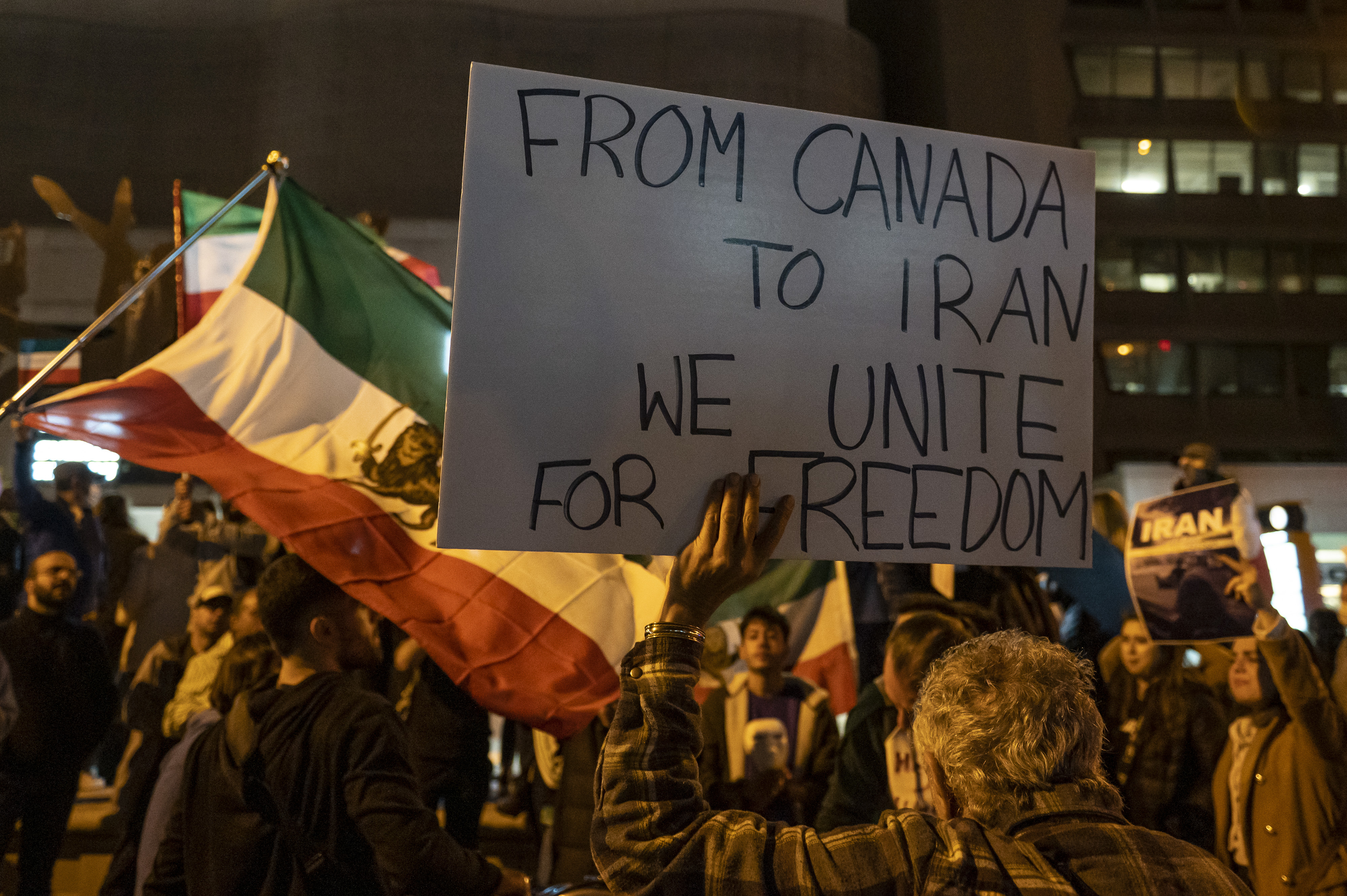 A group of people gather in protest at night in Toronto, some are holding Iranian flags and in the foreground, a man holds a sign that says "From Canada to Iran we unite for freedom" 