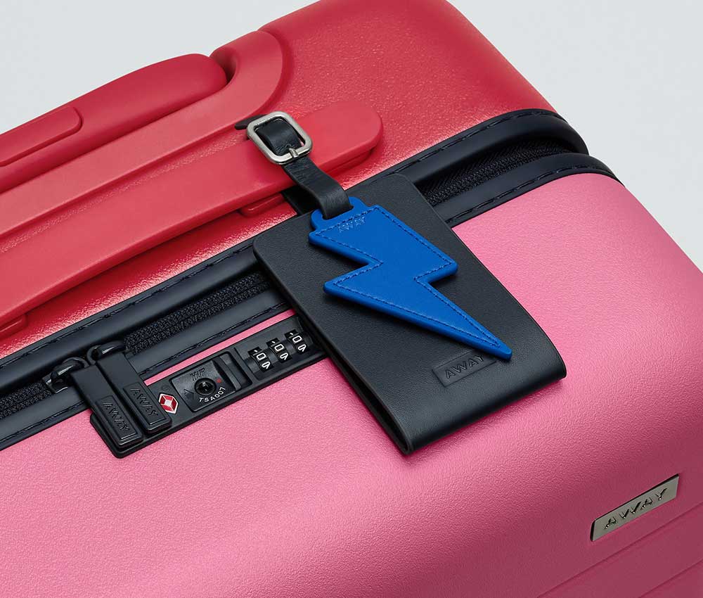 Blue leather Away luggage tag charm shaped like a lightening bolt photographed on a pink and red suitcase.
