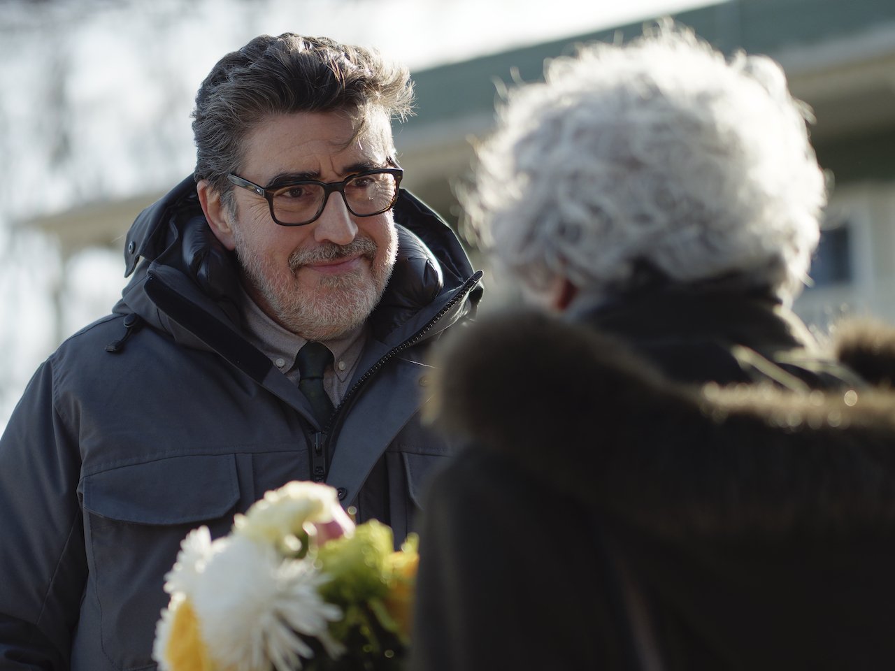A photo of a man with glasses speaking to a woman with white hair. Both are wearing winder coats and the woman is carrying flowers.