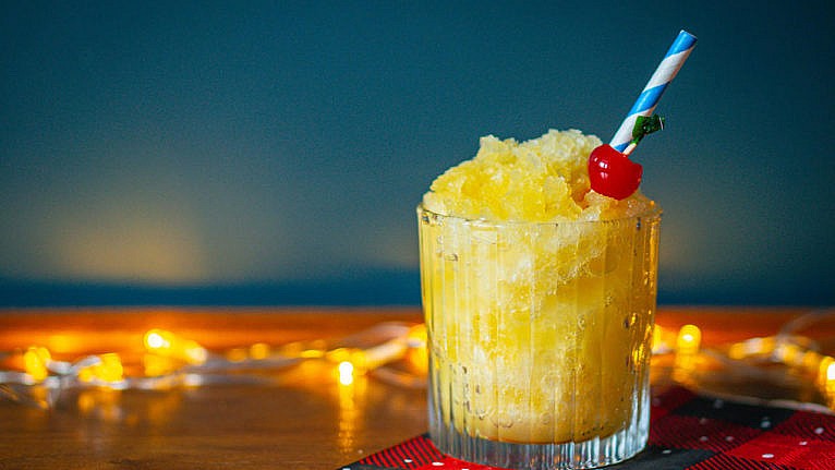 a glass of yellow pineapple slush with a cherry and paper straw
