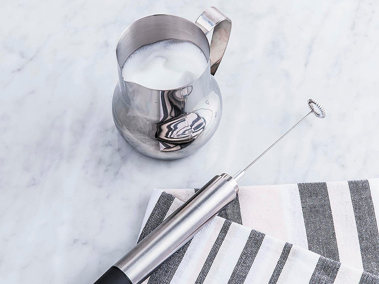 A Kitchen Stuff Plus milk frother shown on a marble countertop with a jar of milk and striped grey linen cloth.