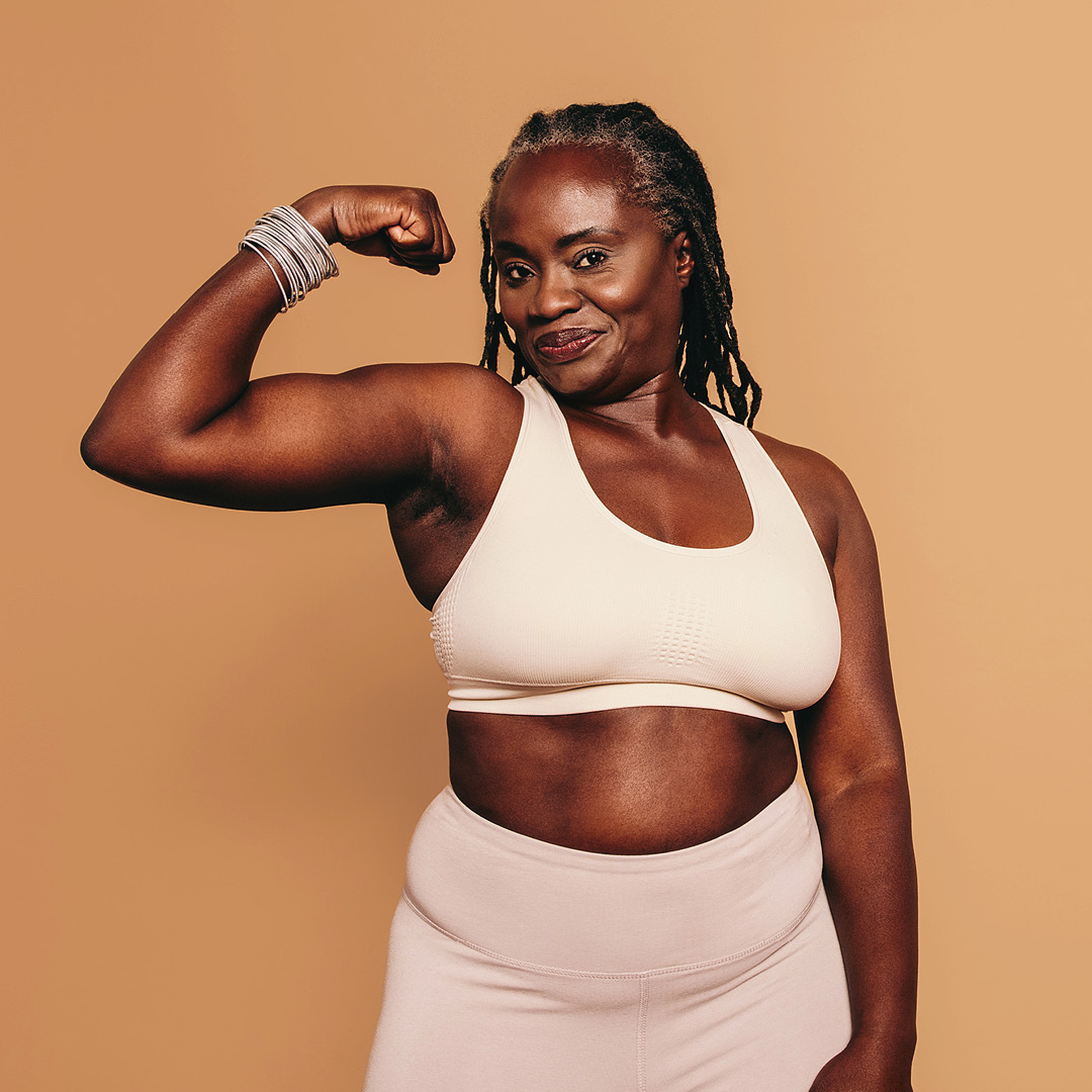 woman in cream coloured leggings and sports bra flexing arm on tan background