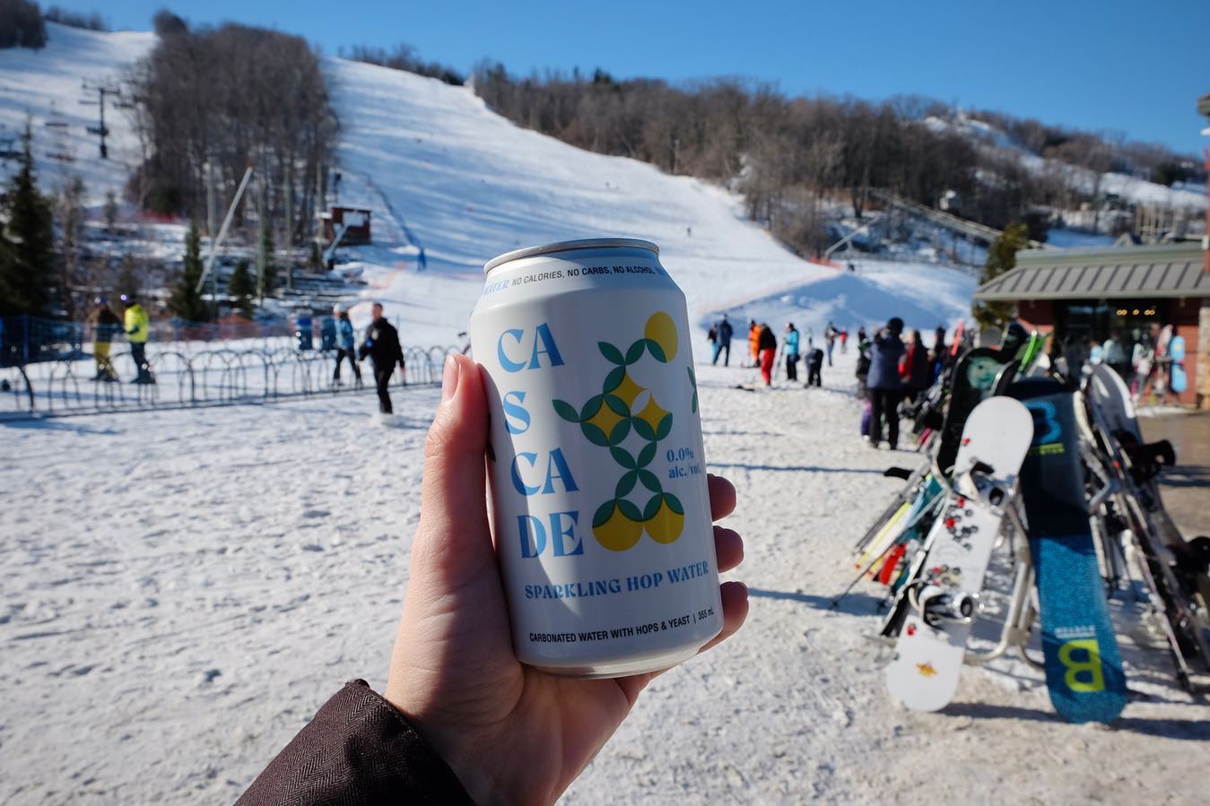 a hand holding a can of cascade sparkling hop water in front of a ski hill