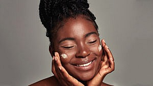 A woman with glowing skin applying cream on both cheeks and smiling.
