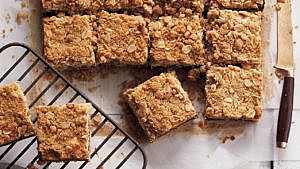 a batch of date squares sitting on parchment paper, freshly cut into squares, with a knife to one side and two squares to the side on a cooling rack