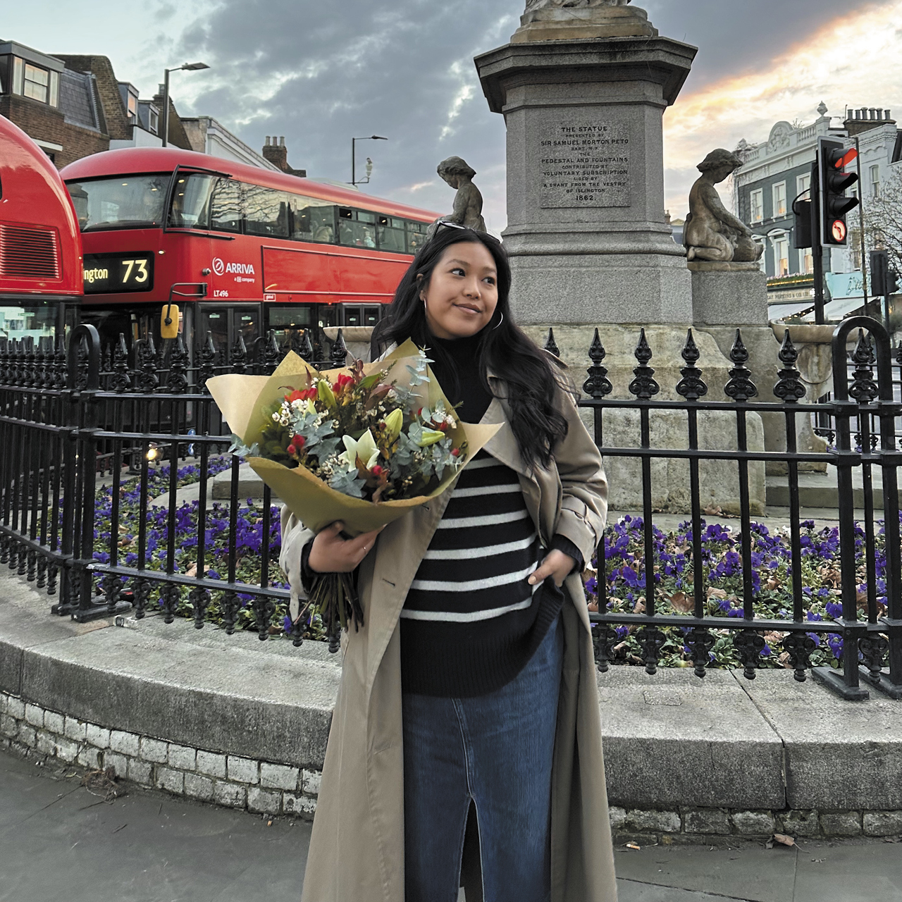 The writer in a striped shirt and trench carrying a bouquet of flowers