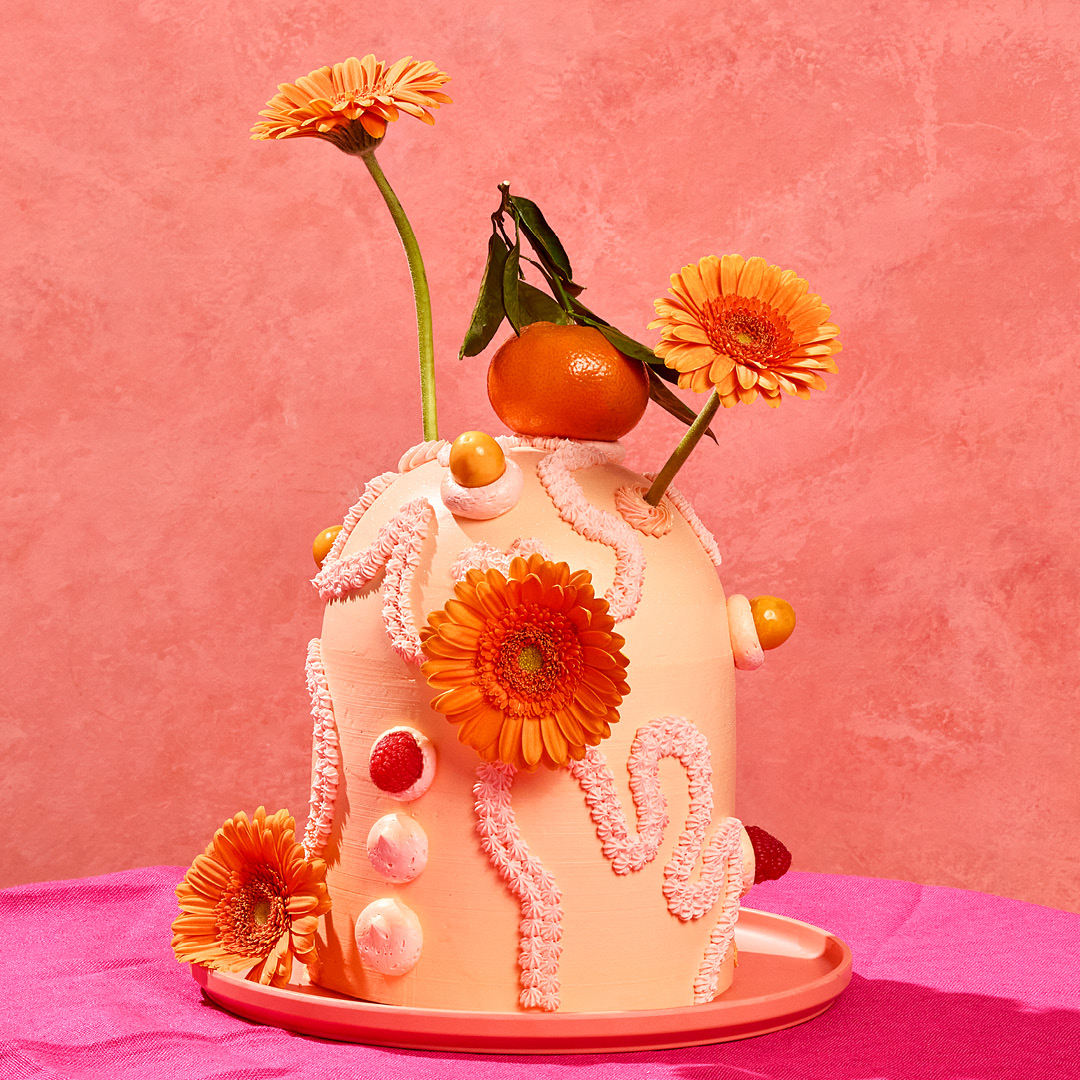 A photo of a tall, domed cake frosted with orange icing and adorned with a clementine and orange gerbera daisies.