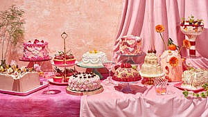 A photo of multiple cakes on platters, on a table draped in pink fabric.