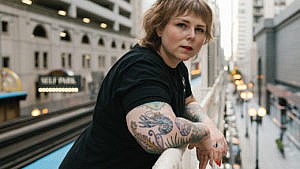 Author Tracey Lindeman leans against a railing in a city, looking at the camera. Lindeman suffered with endometriosis for decades before having a hysterectomy.