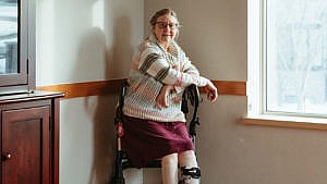 A photo of a woman, sitting on a walker in the corner of a room