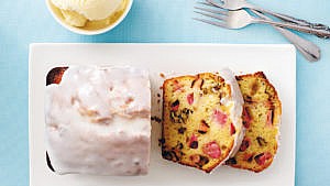 An overhead shot of a Rhubarb-buttermilk tea cake loaf, with white glazing, with two slices cut off it, on a white cutting board on a light blue background with a bowl of vanilla ice cream above it and two forks to one side