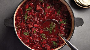 A bowl of beef and beet borscht topped with sprigs of dill with a large silver soup spoon resting in it on a grey table with a bowl of sour cream in the corner