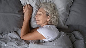 An older woman sleeping in a bed on her left side under a grey blanket