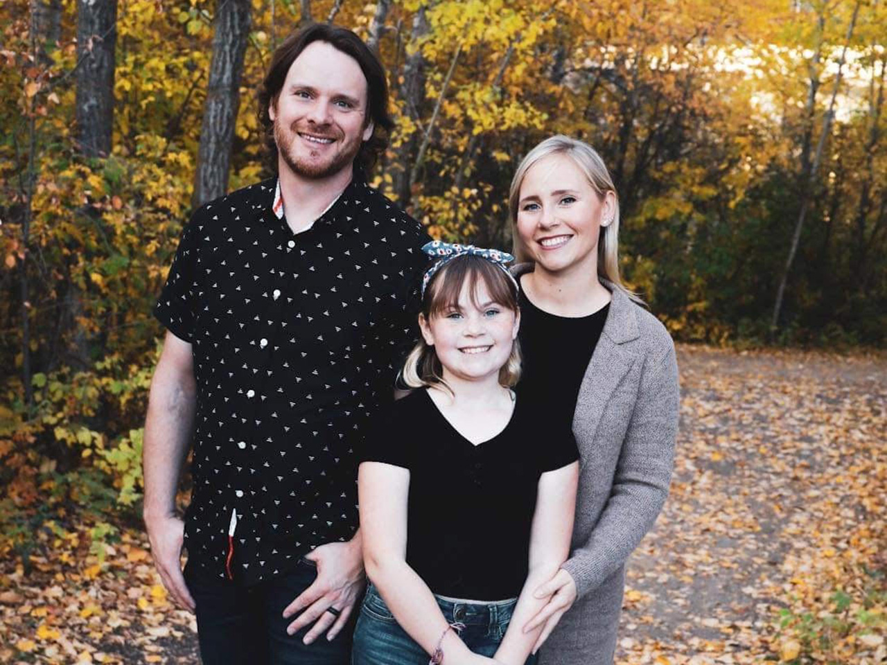 A family of three: from left, a man with brown hair, a beard and black short-sleeved button-down, a young girl wearing a black T-shirt and a woman with blonde hair and a grey cardigan, all standing in a wooded area