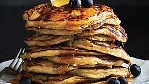 A stack of pancakes on a white plate with a fork topped with a pat of butter and blueberries getting drizzled with maple syrup