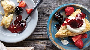 A blue plate with a scone topped with whipped cream and jam with a side of berries on a wood table with another plate with a half-eaten scone and a spoon full of jam