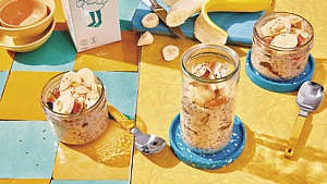 Overnight chia oatmeal in three different mason jars—two short and one tall—on a yellow table beside a sliced banana and yellow spoons and a carton of oat milk