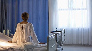A woman with short blonde hair in a white hospital gown sits facing a blue curtain and a window on her hospital bed