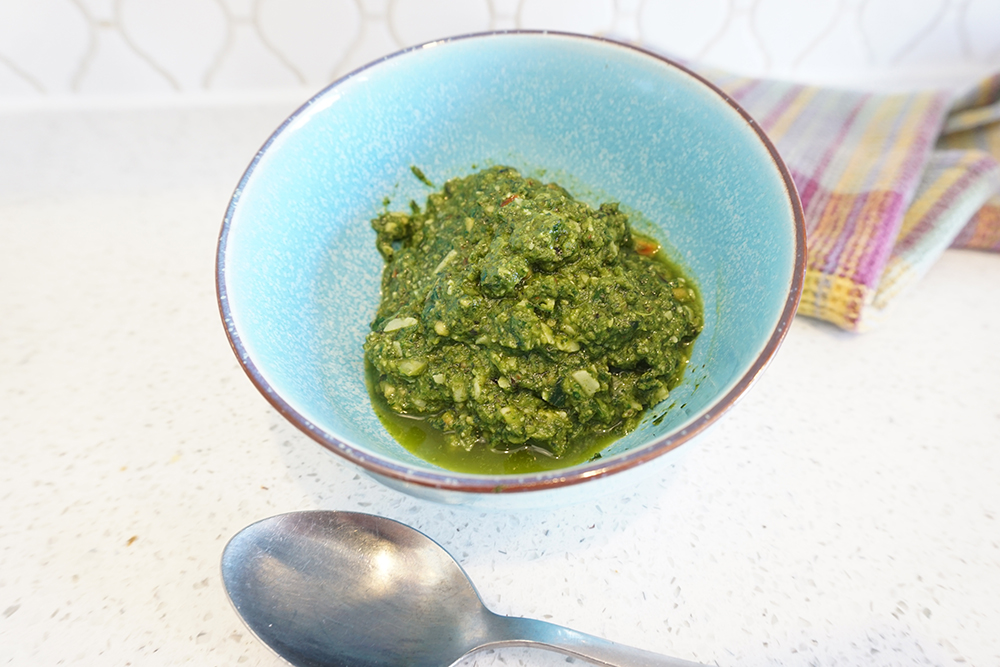 A blue bowl holding spring greens pesto beside a spoon