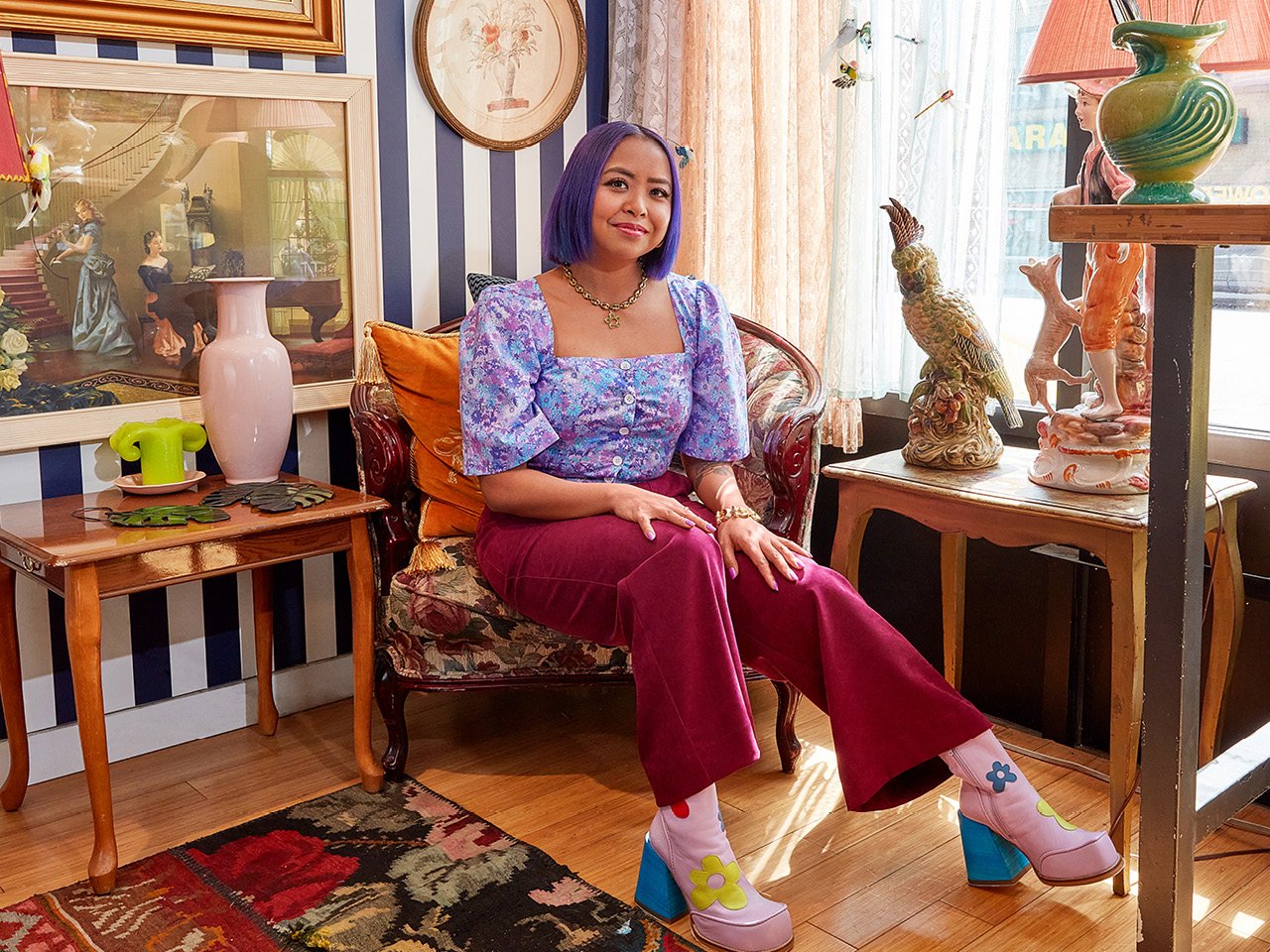 Survivor winner Erika Casupanan poses wearing a short-sleeved purple blouse and red pants, in an armchair against a navy and white striped wall