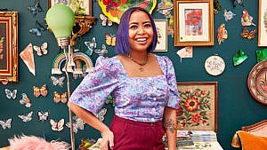 Survivor winner Erika Casupanan, with purple hair and a short-sleeved purple-printed blouse, poses in front of a deep green wall covered with photo frames
